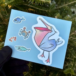 May include: A light blue card featuring stickers of a pelican, fish, and crabs. The pelican sticker is primarily blue with a pink and yellow beak. The fish and crabs are various colors, including blue, orange, and green.