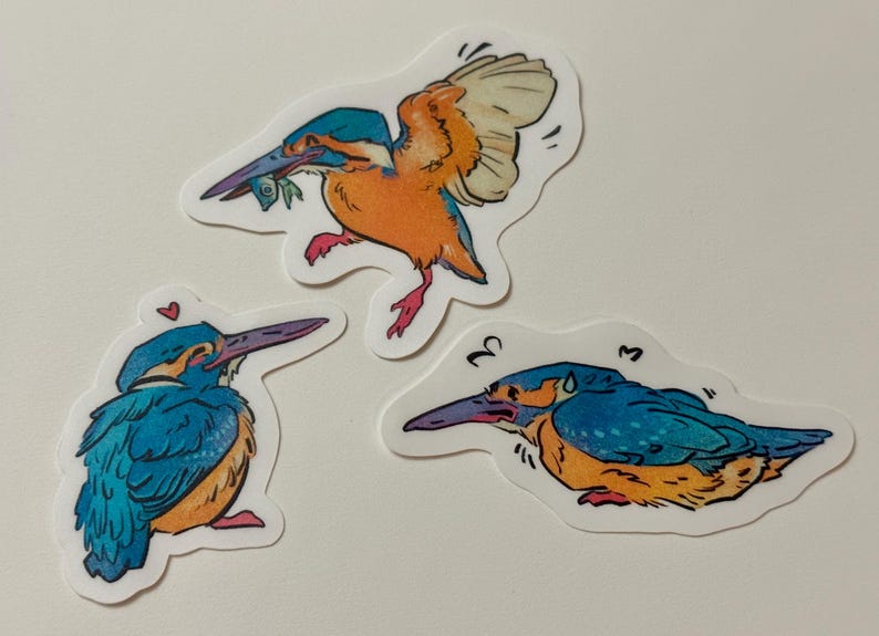 Peut inclure: Trois autocollants repr&eacute;sentant des illustrations de martins-p&ecirc;cheurs. Les oiseaux sont repr&eacute;sent&eacute;s dans diff&eacute;rentes poses, avec des couleurs bleues, oranges et jaunes vives. L'un des oiseaux vole avec un poisson dans le bec, tandis que les autres sont perch&eacute;s.