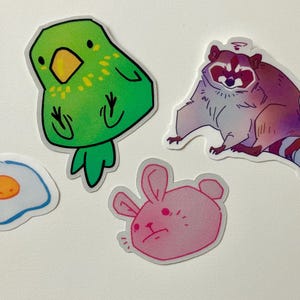 May include: Four colorful stickers featuring a green parrot, a pink bunny, a blue and white egg, and a purple raccoon.