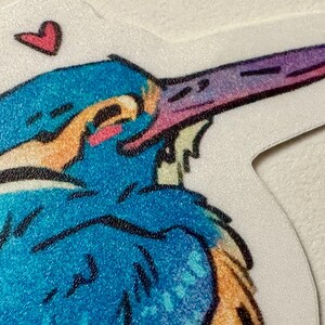 Peut inclure: Un autocollant repr&eacute;sentant une illustration color&eacute;e d'un oiseau au long bec. L'oiseau a des teintes bleues, oranges et violettes, avec un petit c&oelig;ur rose au-dessus de sa t&ecirc;te. L'autocollant a une bordure blanche.