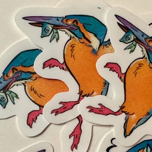 Peut inclure: Plusieurs autocollants repr&eacute;sentant un martin-p&ecirc;cheur stylis&eacute;. L'oiseau a une t&ecirc;te turquoise, un corps orange et des pieds roses, tenant un petit poisson dans son bec. Les autocollants sont blancs avec une bordure d&eacute;coup&eacute;e.