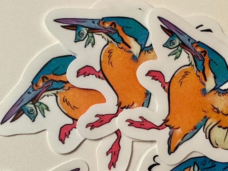 Peut inclure: Plusieurs autocollants repr&eacute;sentant un martin-p&ecirc;cheur stylis&eacute;. L'oiseau a une t&ecirc;te turquoise, un corps orange et des pieds roses, tenant un petit poisson dans son bec. Les autocollants sont blancs avec une bordure d&eacute;coup&eacute;e.