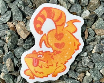 May include: A whimsical sticker featuring an orange and yellow cartoon lizard with a long, striped tail and heart-shaped markings. The lizard has its tongue sticking out and is set against a white background.