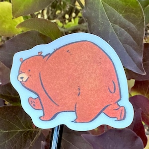 May include: A sticker featuring a cartoon bear in shades of brown and orange, outlined in black, with a light blue border. The bear is depicted walking with a friendly expression. The sticker is likely for decorative purposes.