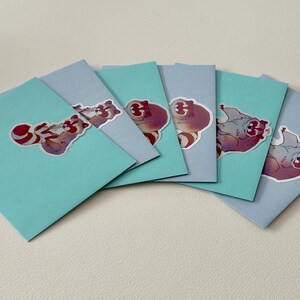 May include: Six light blue cards with colorful cartoon raccoon stickers on them. The stickers feature a raccoon with a red and white striped tail.