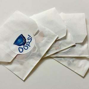 May include: Five white paper envelopes with a sticker on one of the envelopes. The sticker is round and has a blue and white illustration of a broken cup with the word "OOPS!" in blue text.