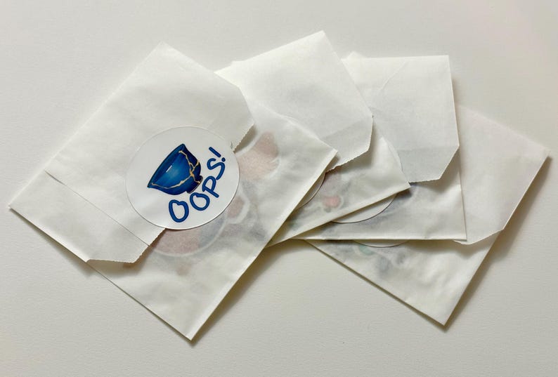 May include: Five white paper envelopes with a sticker on one of the envelopes. The sticker is round and has a blue and white illustration of a broken cup with the word "OOPS!" in blue text.