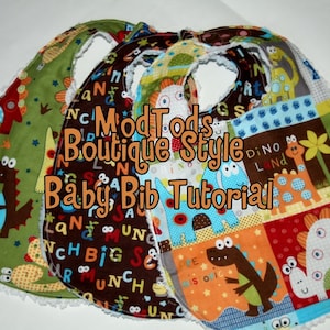 May include: Three handmade baby bibs with dinosaur and alphabet patterns. The bibs feature a variety of colors, including green, brown, blue, and orange. The text "ModTods Boutique Style Baby Bib Tutorial" is displayed on the bibs.