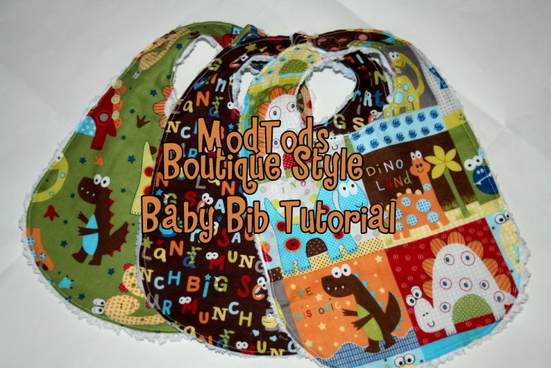 May include: Three handmade baby bibs with dinosaur and alphabet patterns. The bibs feature a variety of colors, including green, brown, blue, and orange. The text "ModTods Boutique Style Baby Bib Tutorial" is displayed on the bibs.