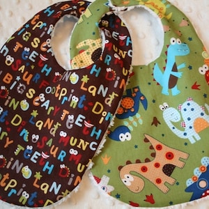 May include: Two baby bibs with dinosaur designs. One bib is brown with colorful text and dinosaur graphics. The other is green with various dinosaur illustrations in blue, orange, and brown. Both have a soft, white backing.