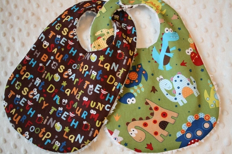 May include: Two baby bibs with dinosaur designs. One bib is brown with colorful text and dinosaur graphics. The other is green with various dinosaur illustrations in blue, orange, and brown. Both have a soft, white backing.