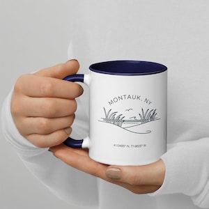 May include: White ceramic coffee mug with a navy blue rim and handle. The mug features a line drawing of a beach scene with grass, a road, and a bird flying over the ocean. The text "Montauk, NY" and "41.0486° N, 71.9605° W" are printed on the mug.
