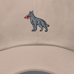 May include: A tan baseball cap with a curved brim. The cap features an embroidered design of a gray dog holding a red object in its mouth. The cap has a six-panel design with a button on top.
