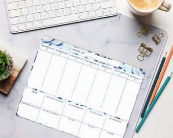 May include: A weekly planner with a blue floral design, featuring sections for each day of the week and additional notes. A white keyboard, pencils, and a cup of coffee are also on the desk.
