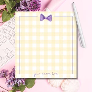 May include: A yellow and white gingham patterned notepad with a purple bow at the top. The notepad has a thin purple border and the words "your name here" at the bottom. A white pen rests on the right side of the notepad.