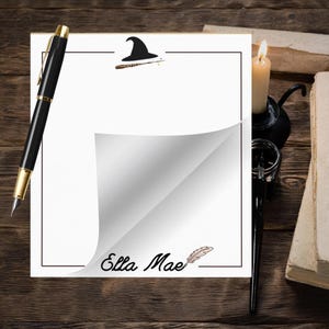 May include: A white notepad with a black border, featuring a curled corner revealing a blank page. The notepad has a black pen with gold accents, a lit candle, and the text "Ella Mae" with a feather illustration. A wizard hat and wand are at the top.