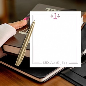 May include: A gold pen rests on a stack of notebooks next to a notepad. The notepad has a pink border and a pink scale of justice icon. The notepad is labeled "Ella Nicole, Esq."