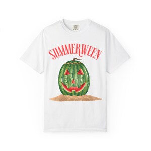 May include: White t-shirt with the word "SUMMERWEEN" in red above a green watermelon carved like a jack-o'-lantern. The watermelon has red accents and sits on a bed of sand.
