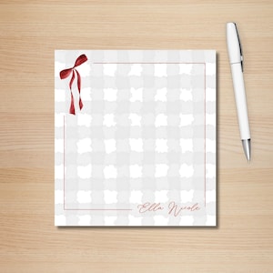 May include: A notepad with a gray and white checkered pattern, a red ribbon, and a red border. The notepad has the name "Ella Nicole" written in red script. A white pen with a silver tip rests on a wooden surface.