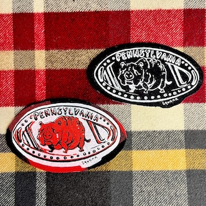 May include: Two oval patches featuring the text "PENNSYLVANIA WILD" and a pig illustration. One patch is red and white, the other is black and white. They are set against a red, black, and yellow plaid background. The word "Squonk" is written on the bottom of each patch.