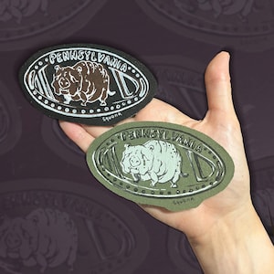 May include: Two oval-shaped embroidered patches featuring a creature illustration and the text "PENNSYLVANIA" and "Squonk". One patch is black with a brown creature, and the other is olive green with a light green creature, both with white outlines.