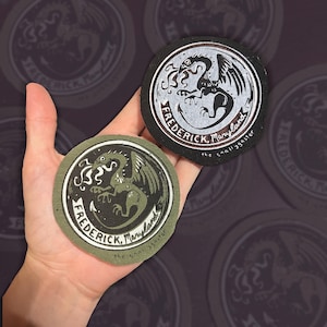 May include: Two circular embroidered patches featuring a mythical creature design. One patch is black with white details, the other is olive green with black details. Both display the text "FREDERICK, Maryland" and "the snallygaster".
