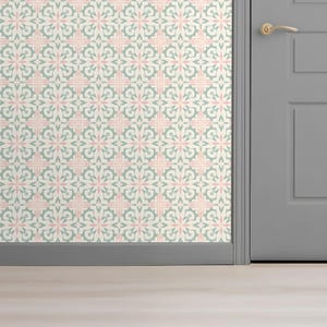 Może przedstawiać: Fragment ściany pokryty wzorzystą tapetą. Tapeta przedstawia powtarzający się kwiatowy wzór w miękkim różu i zieleni na kremowym tle. Szare drzwi i listwy są widoczne po prawej stronie, ze złotą klamką.