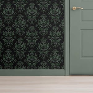 Op de afbeelding: Een hoek van een kamer met zwart behang met groene bloemmotieven. Een gedempte groene deur met een gouden handvat is zichtbaar, samen met een bijpassende groene plint. De vloer is lichtgekleurd hout.
