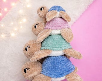 Peut inclure: Une pile de tortues crochetées bleues, vertes et roses, avec le texte "PATTERNS TORTUES" et "Luna Olivia Crochet". Les tortues ont un aspect doux et texturé. Le fond est rose et blanc, avec des guirlandes lumineuses.