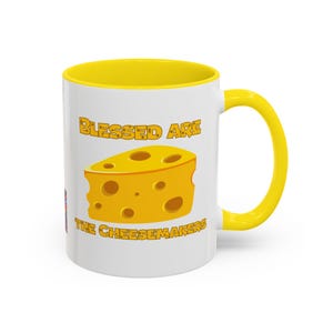 May include: White ceramic mug with a yellow interior and handle. The mug features the text "BLESSED ARE" above a cartoon cheese wedge and the text "THE CHEESEMAKERS" below. The cheese wedge is yellow with several holes.