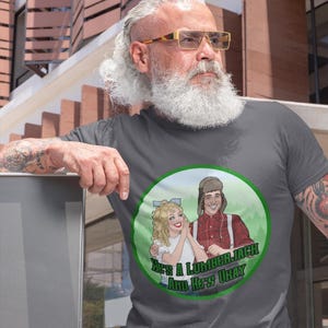May include: A gray t-shirt featuring a circular graphic with the text "He's a Lumberjack and He's Okay." The graphic depicts a cartoon couple in a forest setting. The man is wearing a red plaid shirt and the woman is wearing a white top.