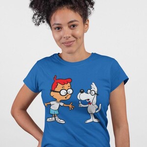 May include: Blue t-shirt featuring a cartoon illustration of a boy and a dog, both wearing glasses. The boy has red hair and is wearing a white shirt and blue shorts. The dog is gray and wearing a red bow tie.