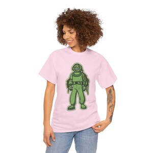 May include: A light pink t-shirt featuring a cartoon illustration of a green diving suit. The diver has a helmet and is wearing a full suit. The illustration is centered on the shirt.