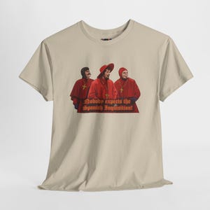 Puede incluir: Camiseta beige con un diseño de tres figuras con túnicas y sombreros rojos, con cruces doradas. El texto "Nobody expects the Spanish Inquisition!" está impreso debajo en rojo y dorado.