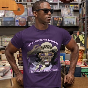 May include: A purple t-shirt featuring a cartoon illustration of a character wearing a hat and sunglasses. The text "IT'S A PIMP NAMED SLICKBACK" is above the image, and "YOU SAY THE WHOLE THING!" is below.