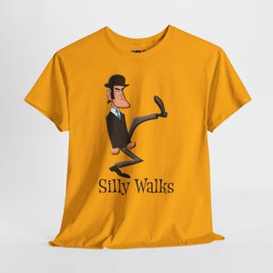 Könnte beinhalten: Senfgelbes T-Shirt mit einer Cartoon-Illustration eines Mannes in einem schwarzen Anzug und einer Melone, der eine "Silly Walks"-Pose ausführt. Der Text "Silly Walks" ist unter der Illustration in schwarzer Schrift gedruckt.