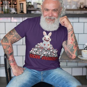 May include: Purple t-shirt featuring a menacing white rabbit with red eyes and blood, sitting atop a pile of skulls. The text "RUN AWAY!" is printed in red below the graphic. The shirt is worn by a person.