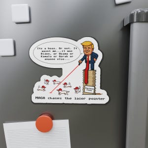 Puede incluir: Un imán blanco con una imagen de dibujos animados de un hombre con un puntero láser, sobre una pila de libros "Epstein Files". El texto dice "MAGA chases the laser pointer" y una burbuja.