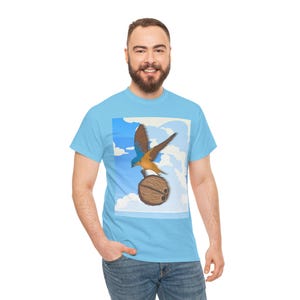 May include: Light blue t-shirt featuring a graphic of a blue and brown bird perched on a coconut against a sky background. The shirt is made of a soft material and is suitable for casual wear.