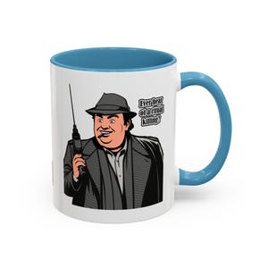 May include: White ceramic mug with a light blue interior and handle. The mug features a cartoon illustration of a man in a fedora and trench coat, holding a gun. Text on the mug reads, "Ever hear of a ritual killing?"