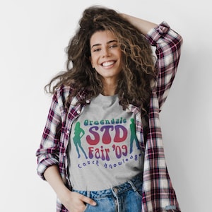 May include: A woman wearing a gray t-shirt with the text "Greendale STD Fair '09" in colorful lettering. The shirt also features silhouette figures. She is wearing a red, white, and black plaid shirt and blue jeans.