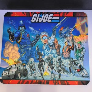 May include: A rectangular mouse pad featuring a colourful G.I. JOE graphic illustration. The artwork depicts a group of characters in tactical gear, a blue-masked figure, military vehicles, and explosions against a blue sky, with the G.I. JOE logo at the top.