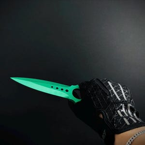 May include: A bright green throwing knife with a pointed blade and four circular cutouts along the centre, held by a black fingerless glove featuring a textured geometric pattern and silver reflective accents on the back of the hand.