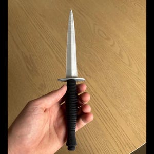 Peut inclure: Un poignard argenté avec une lame pointue et un manche noir texturé. Le poignard a une garde circulaire et une lame effilée. La poignée semble être faite d'un matériau nervuré.