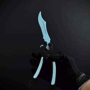 May include: A butterfly knife featuring a light blue curved blade and a black handle with light blue accents. The knife is shown in an open position with a visible pivot mechanism and a small latch near the base of the blade.