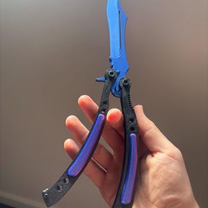 Könnte beinhalten: Ein blau-schwarzes Butterfly-Messer mit gebogener Klinge. Der Griff ist schwarz mit lila und rosa Akzenten. Das Messer wird offen gehalten und zeigt sein Design.