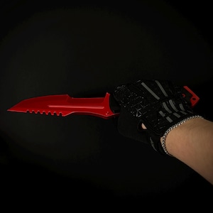 Könnte beinhalten: Ein leuchtend rotes Messer mit feststehender Klinge und gezackter Schneide nahe dem Griff, gehalten von einem schwarzen fingerlosen Handschuh mit geometrischem Muster und grauen Akzenten. Das Messer hat eine Spitze und einen Mittelgrat.