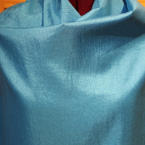 May include: A draped, turquoise-colored fabric, possibly silk or a similar material, is displayed on a dress form. The fabric has a subtle sheen and a textured surface, creating soft folds and shadows. The overall effect is elegant and luxurious.