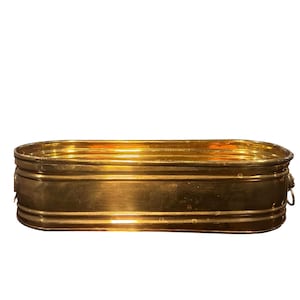 May include: A rectangular brass planter with rounded corners. The planter has a shiny, golden finish and features a series of horizontal bands around its exterior. Small, decorative handles are visible on the sides. The planter is designed for decorative use.