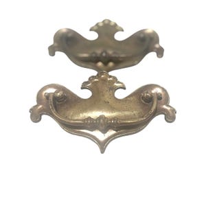 May include: Two antique brass drawer pulls with ornate designs. The pulls have a curved handle and decorative details, including a central flourish and rounded end pieces. The brass has a warm, aged patina, suggesting vintage or antique hardware.